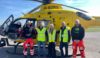 A group of people standing in front of a helicopter. Two of them are air ambulance crew members and three of them are visitors.