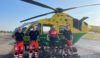 A group of people standing in front of an air ambulance helicopter. Three of the are clinical crew and one is a cyclist, who is standing with his bicycle.