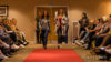 Two ladies in a room walking down a red carpet with people sat watching
